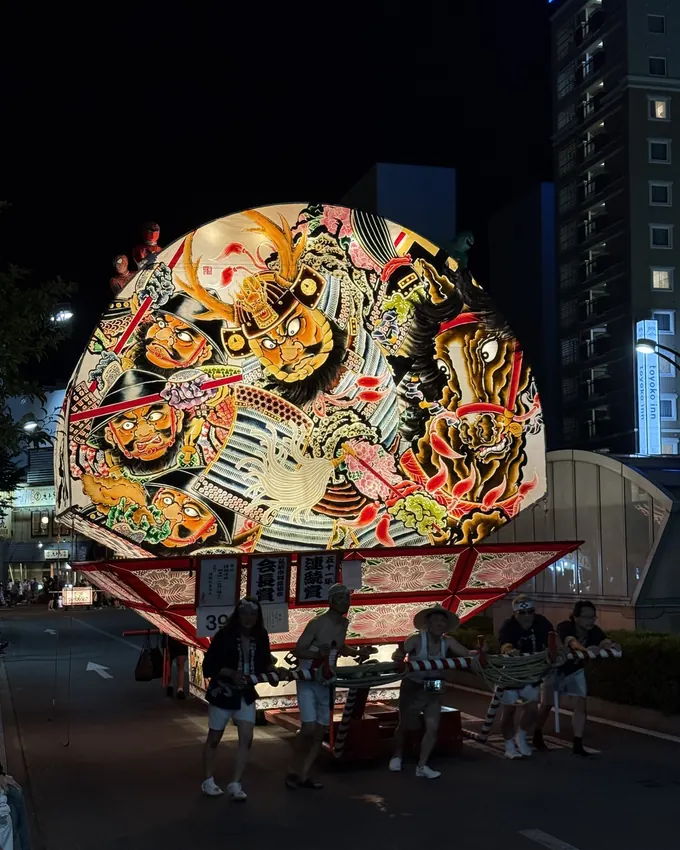 Glowing fan-shaped Neputa float and its pullers on a night street, Hirosaki